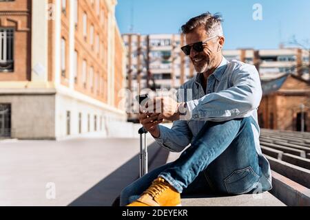 Stock Foto von einem Geschäftsmann in einer Bank auf der Straße sitzen Blick auf sein Handy und Musik hören mit seinen kabellosen Ohrhörern. Er ist ho Stockfoto