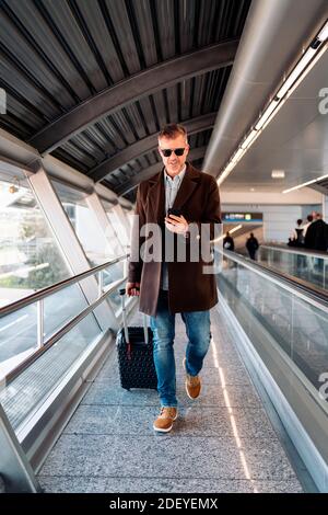 Stock Foto von einem kaukasischen Geschäftsmann zu Fuß in der Nähe eines Förderbandes mit einem Koffer gehen, um einen Flug zu nehmen. Er lächelt und schaut auf sein smartpho Stockfoto