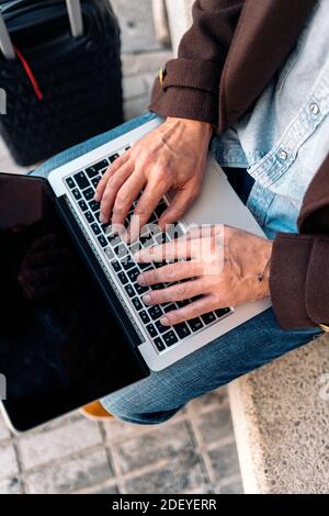 Stock Foto von oben Nahaufnahme eines unkenntlichen Geschäftsmanns, der in seinem Laptop tippt. Er sitzt auf einer Bank. Er hat einen Koffer in seiner Nähe. Stockfoto