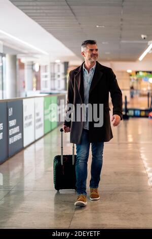 Stock Foto von einem kaukasischen Geschäftsmann, der mit einem Koffer auf dem Flughafen läuft, um einen Flug zu nehmen. Er lächelt und schaut zur Seite. Er ist abgenutzt Stockfoto