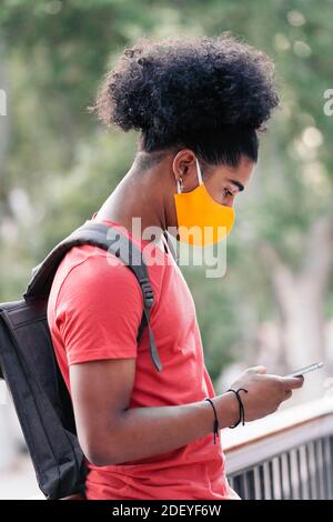 Stock Foto von jungen schwarzen Jungen mit seinem Handy auf der Straße. Er trägt eine Gesichtsmaske wegen covid19. Stockfoto