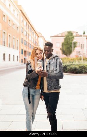 Multiethnisches junges Paar, das mit dem Telefon auf der Straße läuft. Stockfoto