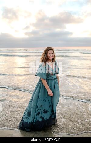 Wunderschöne Frau trägt Maxi-Kleid wating im Meer surfen Bei Sonnenuntergang Stockfoto