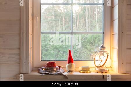 Weihnachtslaterne, Weihnachtsgnome und roter Becher auf dem Fenster eines Holzhauses mit Blick auf den Wintergarten. Stockfoto