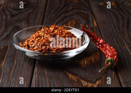 Rote Paprikaflocken in einer Glasuntertasse und trocken zerknittert Pod auf einem dunklen Holztisch. Natürliche Gewürze und Gewürze für Fleisch-, Fisch- und Gemüsegarten. Stockfoto