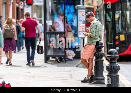 London, Großbritannien - 23. Juni 2018: Junger Mann, der mit dem Telefon auf der Bürgersteig-Straße durch Doppeldecker-Bushaltestelle mit Menschen im Hintergrund in Chelsea, United K stöbert Stockfoto