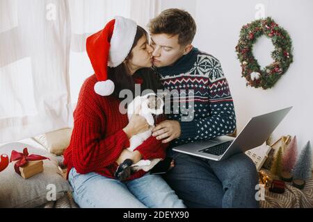 Glückliche junge Familie küssen und halten Kätzchen und Laptop in festlich dekorierten Raum. Stilvolles Paar in Pullover und weihnachtsmütze feiern Weihnachten an Stockfoto