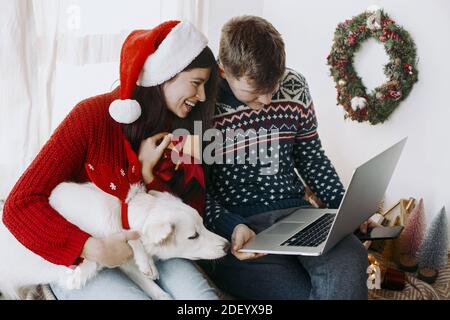 Stilvolles glückliches Paar mit niedlichen Hund Austausch weihnachtsgeschenke mit Familie auf Videoanruf, feiern Urlaub zu Hause. Frohe Feiertage! Junge Familie Stockfoto