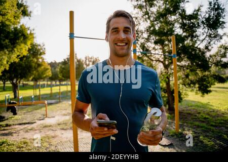 Hübscher junger männlicher Athlet, der im Freien Sport macht und Musik hört Ohrhörer mit Smartphone und Wasserflasche Stockfoto