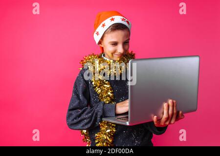 Ein schöner Teenager im Hut des Weihnachtsmannes und Lametta am Hals, mit einem Laptop in den Händen, der Kerl sendet SMS-Grüße. Stockfoto