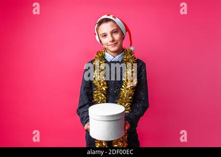 Teen in Santa Hut und mit Lametta am Hals halten Geschenkbox auf rotem Hintergrund. Das Konzept der Weihnachten, um Geschenke zu geben Stockfoto