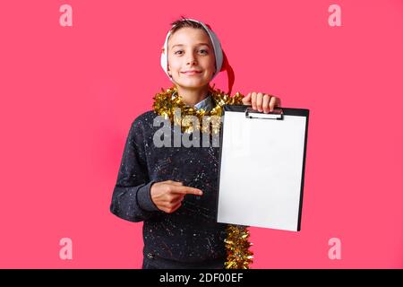 Teen Kerl trägt Weihnachtsmütze, und mit Lametta um den Hals, hält eine Tablette mit einem sauberen Laken und zeigt einen Finger auf ihn, plant das Kommen Stockfoto