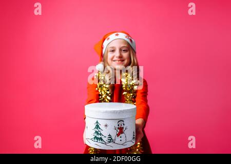 Teen Mädchen in Weihnachtsmann Weihnachtsmütze und Lametta um ihren Hals, gibt ein Weihnachtsgeschenk auf einem roten Hintergrund. Das Konzept der Weihnachten, um Geschenke zu geben Stockfoto