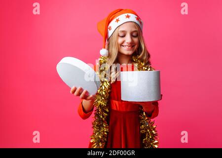 Fröhliches Mädchen in Weihnachtsmütze und mit Lametta am Hals guckt in Geschenkbox für Weihnachten auf rotem Hintergrund. Stockfoto