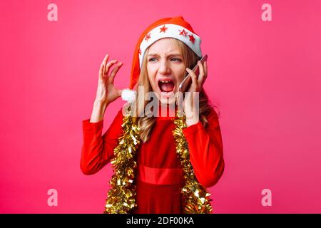 Porträt eines wütenden Schulmädchen in Weihnachtsmütze und Lametta am Hals, emotional am Telefon sprechend und schreiend, isoliert auf rotem Hintergrund Stockfoto