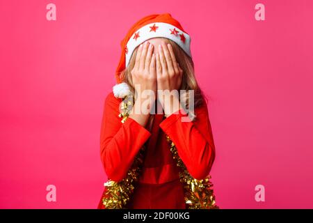 Mädchen im roten Hut des Weihnachtsmannes und mit Lametta am Hals, schließt ihre Augen mit den Händen und versteckt sich vor jemandem auf rotem Hintergrund Stockfoto