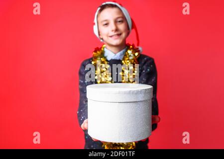 Ein Teenager in einem Weihnachtsmütze und Lametta auf dem Hals, gibt ein Weihnachtsgeschenk, auf einem roten Hintergrund. Das Konzept der Weihnachten, um Geschenke zu geben Stockfoto