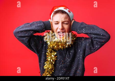 Porträt eines Teenagers in einem Weihnachtsmann Hut und Lametta auf dem Hals, schreiend und bedeckt seine Ohren Kerl, will nichts hören. Weihnachten, menschliche Emotionen, Stockfoto