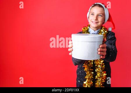 Ein Teenager in einem Weihnachtsmütze und Lametta auf dem Hals, gibt ein Weihnachtsgeschenk, auf einem roten Hintergrund. Das Konzept der Weihnachten, um Geschenke zu geben Stockfoto