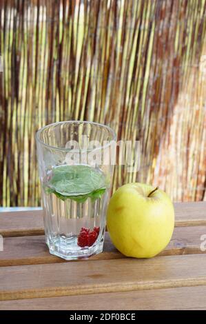 Ein flacher Fokus vertikale Aufnahme von grünen Blättern und Himbeere Obst in einem Glas Wasser auf einem Holztisch Mit einem gelben Apfel an der Seite Stockfoto