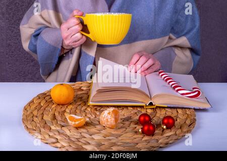 Nahaufnahme Stillleben mit einem Buch, Weihnachtsdekor auf dem Tisch. Eine Frau mittleren Alters in einer Stola hält eine große gelbe Tasse heißes Getränk. Selektiver Fokus. Stockfoto