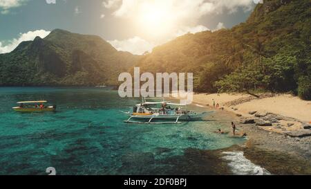 Sonne über Bergketten mit grünem Tropenwald an der Ocean Bay. Die Menschen ruhen am Sandstrand in der Nähe von Wanderbooten. Tourist Sommerurlaub an sonnigen Tag auf Palawan Island, Philippinen, Asien. Luftaufnahme Stockfoto