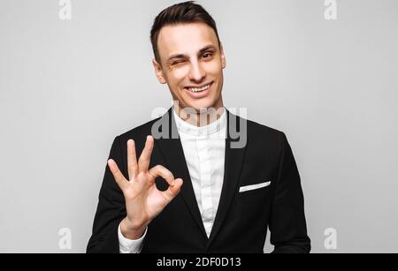 Porträt eines jungen, gutaussehenden glücklichen Mannes, in einem Geschäftsanzug, lächelnd und mit einer OK-Geste, auf grauem Hintergrund. Positive menschliche Emotionen, Gesten Stockfoto