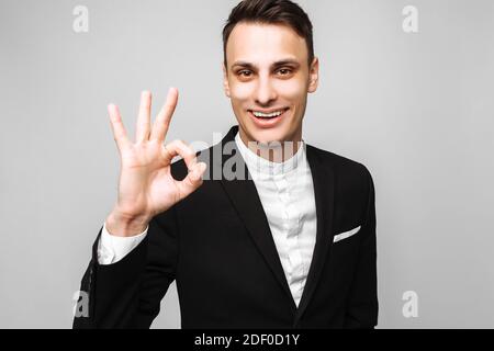 Porträt eines jungen, gutaussehenden glücklichen Mannes, in einem Geschäftsanzug, lächelnd und mit einer OK-Geste, auf grauem Hintergrund. Positive menschliche Emotionen, Gesten Stockfoto
