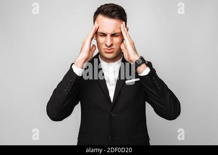 Schöner junger Geschäftsmann, männlich, in einem klassischen schwarzen Anzug, fühlt sich starke Kopfschmerzen, vor einem grauen Hintergrund. Stockfoto