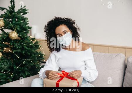Junge afroamerikanische Frau trägt Gesichtsmaske und hält eine Geschenkbox zu Hause in dekorierten Raum, Online-Weihnachtseinkäufe und Geschenk-Schenken per Post Stockfoto
