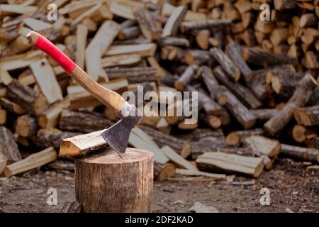 Eine hölzerne, alte Axt in ein gesägtes, altes Stück Holz vor dem Hintergrund des gehackten Brennholzes eingefügt. Stockfoto