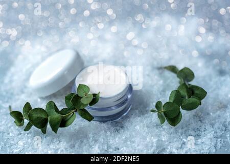 Kosmetische Creme in weißem Glas für Gesicht und Hand Hautpflege. Weißer schneebedeckter Hintergrund, Eukalyptusblätter. Konzept der Winterpflege. Stockfoto