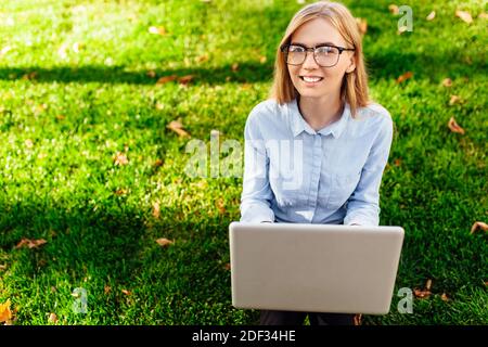 Junge attraktive Mädchen mit Brille, Frau auf dem Gras sitzen, auf einem Laptop arbeiten, in einem Stadtpark auf einem grünen Rasen im Freien. Freiberuflicher Geschäftsbetrieb Stockfoto