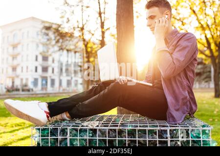 Der Mann sitzt in legerer Kleidung auf einer Bank, arbeitet mit einem Laptop und hört Musik mit kabellosen Kopfhörern vor dem Hintergrund des Sonnenuntergangs Stockfoto