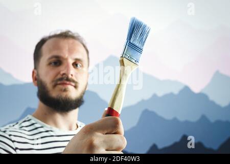 Bärtiger Malermaler, der den Pinsel vor der Wand hält. Wie interessant ist es, die Wand zu bemalen Stockfoto