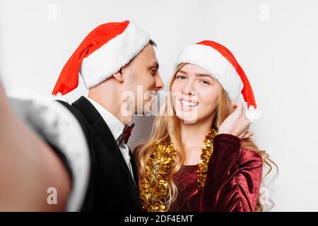 Glückliches Paar von Liebenden in Weihnachtsmannmützen, küsst der Kerl das Mädchen und macht ein Foto am Telefon, mit Weihnachtsgeschenken in ihren Händen. Auf einer weißen BA Stockfoto