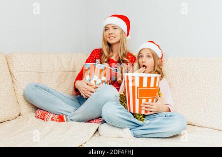 Eine junge Mutter und ihre kleine Tochter, in Weihnachtsmannmützen gekleidet, schauen Filme und essen Popcorn, während sie zu Hause auf der Couch sitzen. Christus Stockfoto