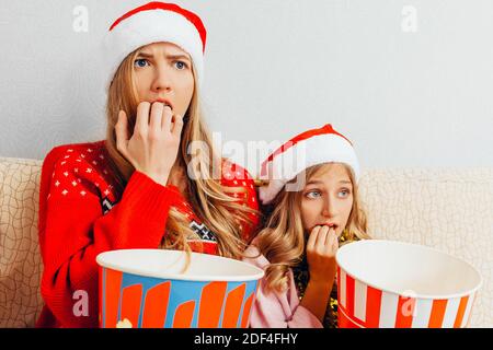 Betäubt Mutter und ihre kleine Tochter, gekleidet in Weihnachtsmannmützen, schauen Filme und essen Popcorn, sitzen auf der Couch. Weihnachtskonzept Stockfoto