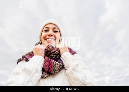 Glücklich junge kaukasische Frau Blick auf die Kamera trägt weiße Wollmütze, gestrickte Zöpfe Pullover mit farbigem Schal in Winterfarben - Portrait of si Stockfoto