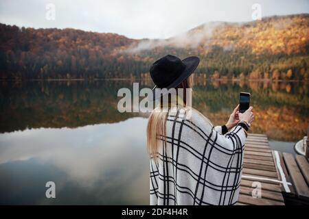 Hipster Frau fotografiert Sonnenuntergang auf dem Smartphone. Mädchen hält ein Handy. Kamera-App. Schöner Herbstabend in der Nähe des Sees. Stockfoto