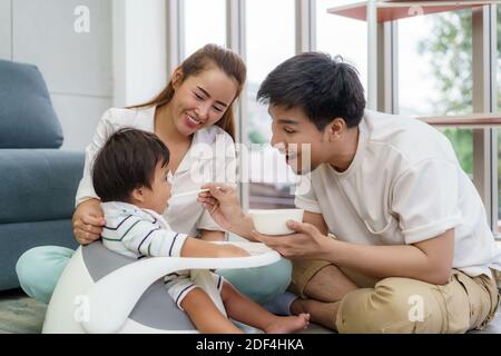Asiatische Vater Fütterung seiner 6 Monate alten Baby Junge mit festen Lebensmitteln mit Löffel und Mutter sitzt in der Nähe, um ihren Sohn zu essen im Wohnzimmer zu Hause jubeln. Stockfoto