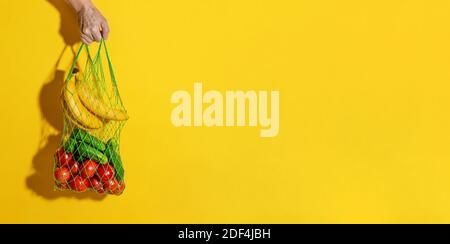 Männliche Hand hält einen weißen Netzbeutel mit Gemüse auf gelbem Hintergrund mit Kopierraum. Zero Waste Shopping-Konzept. Langes, breites Banner Stockfoto