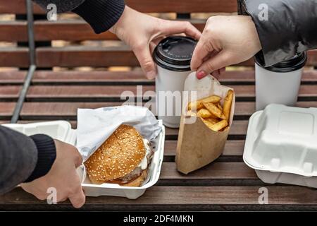 Weibliche Hände legen Hamburger mit Tassen Kaffee, Pommes auf einer Parkbank. Fast Food. Essen zum Mitnehmen Konzept. Stockfoto