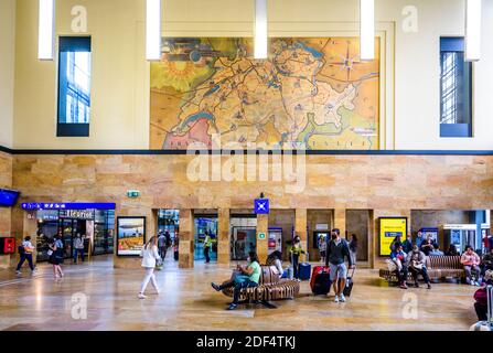 Die Passagiere warten, sitzen auf Bänken oder ziehen rollende Gepäckwagen in der großen historischen Bahnhofsanlage des Genfer Cornavin-Bahnhofs. Stockfoto