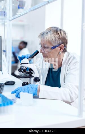 Senior Scientist Frau mit Schutzbrille tun genetische Untersuchung mit Mikroskop mit afrikanischen Assistenten im Hintergrund.. Ältere Forscher, die wissenschaftliche Forschung in einem sterilen Labor mit einer modernen Technologie. Stockfoto