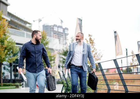 Zufriedene Kollegen auf dem Weg ins Büro diskutieren über das Projekt. Geschäftsleute, die draußen gehen, Papiere halten, reden, lachen. Stockfoto