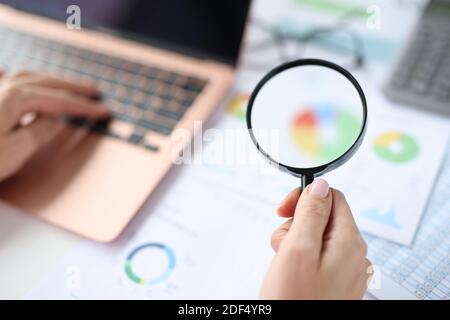 Frau hält Lupe in Händen über Dokumente mit Grafiken Und Diagramme und Arbeiten am Laptop Stockfoto