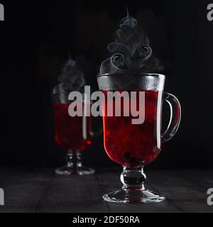 Weihnachts Glühwein - heißer roter Beerentee mit Weihnachtsbaumform Dampf in zwei Glas Weingläser mit reifen Beeren in dunkel eleganten schwarzen Innenraum, Stockfoto