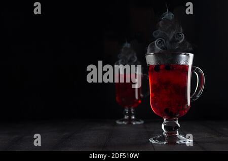 Weihnachtlicher roter Glühwein mit Beeren und Dampf als Weihnachtsbaumform in zwei transparenten Weingläsern auf schwarzem Holztisch im dunklen Innenraum, Cop Stockfoto