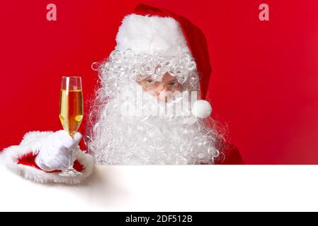 Weihnachtsmann hält ein Glas Sekt in der Hand, während er hinter dem weißen Brett steht. Stockfoto
